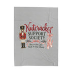 A light gray fabric item, likely a towel or blanket, featuring a Nutcracker-themed graphic print with a nutcracker soldier, ballet slippers, and the text 'Nutcracker SUPPORT SOCIETY Not in the Cast Still in the Chaos'.