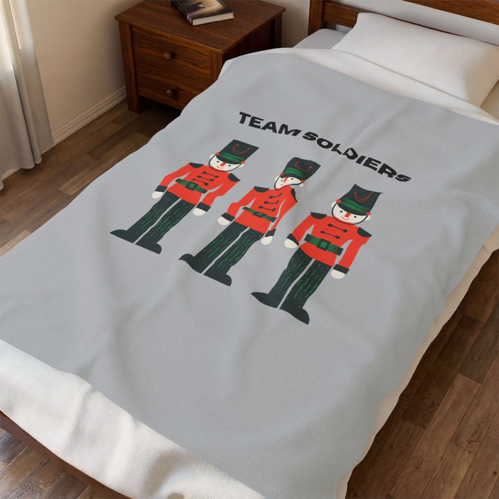 A light gray blanket with a 'TEAM SOLDIERS' graphic featuring three toy soldier illustrations, shown on a bed in a bedroom setting.