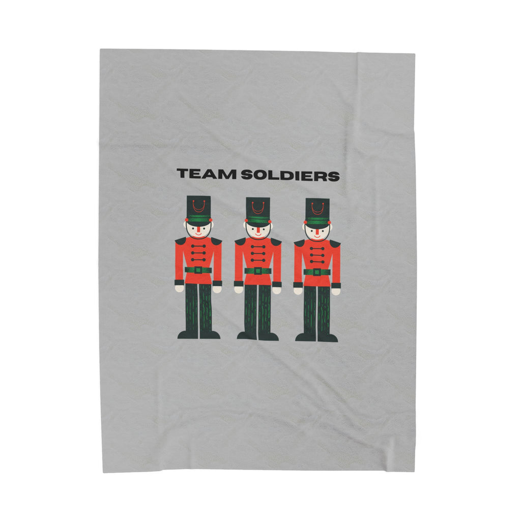 A light gray fabric item, likely a towel or blanket, featuring a graphic print of three toy soldier nutcrackers and the text 'TEAM SOLDIERS' in black.