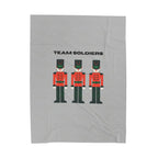 A light gray fabric item, likely a towel or blanket, featuring a graphic print of three toy soldier nutcrackers and the text 'TEAM SOLDIERS' in black.