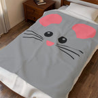 A gray blanket with a cartoon mouse face design, featuring pink ears, black whiskers, and eyes, shown on a bed in a bedroom setting.