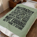 A sage green blanket with a black typographic design featuring the text 'NUTCRACKER NOT KIDDING WE' DONE IT BEFORE' is draped over a bed in a bedroom setting.