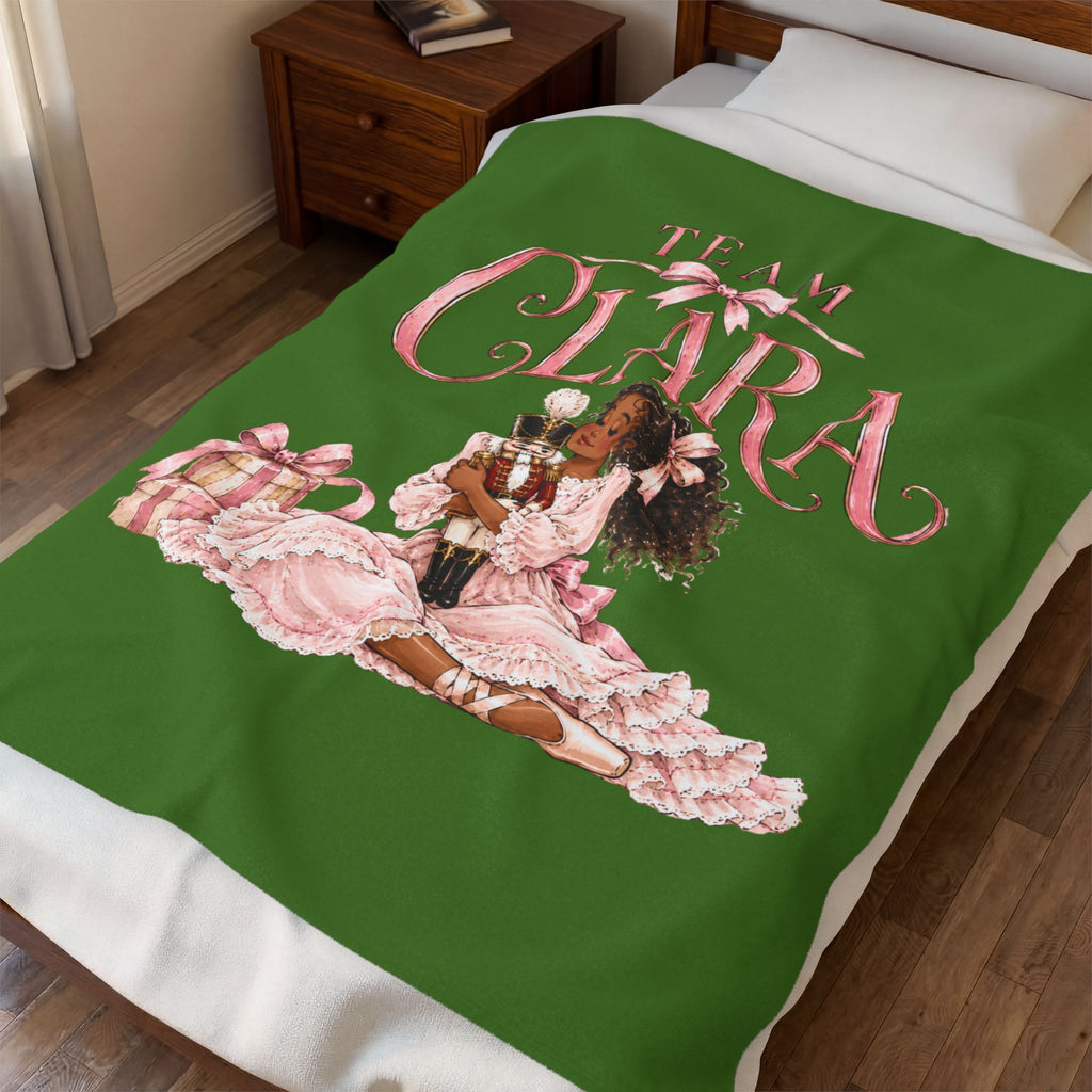 A green blanket with a pink and white graphic of a woman in a ruffled dress and the text 'TEAM CLARA' is shown on a bed in a bedroom setting.