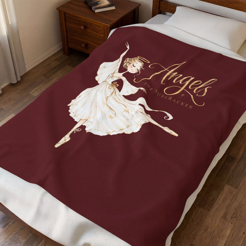 A burgundy blanket with a gold and white illustration of an angel ballerina and the text 'Angels Are Nutcrackers' is shown on a bed in a bedroom setting.