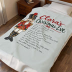 A personalized Christmas blanket laid out on a bed in a bedroom. The blanket is off-white and features a large printed design with a nutcracker illustration, red and green script text reading 'Clara's Christmas Eve,' and a list of holiday song titles in black text.