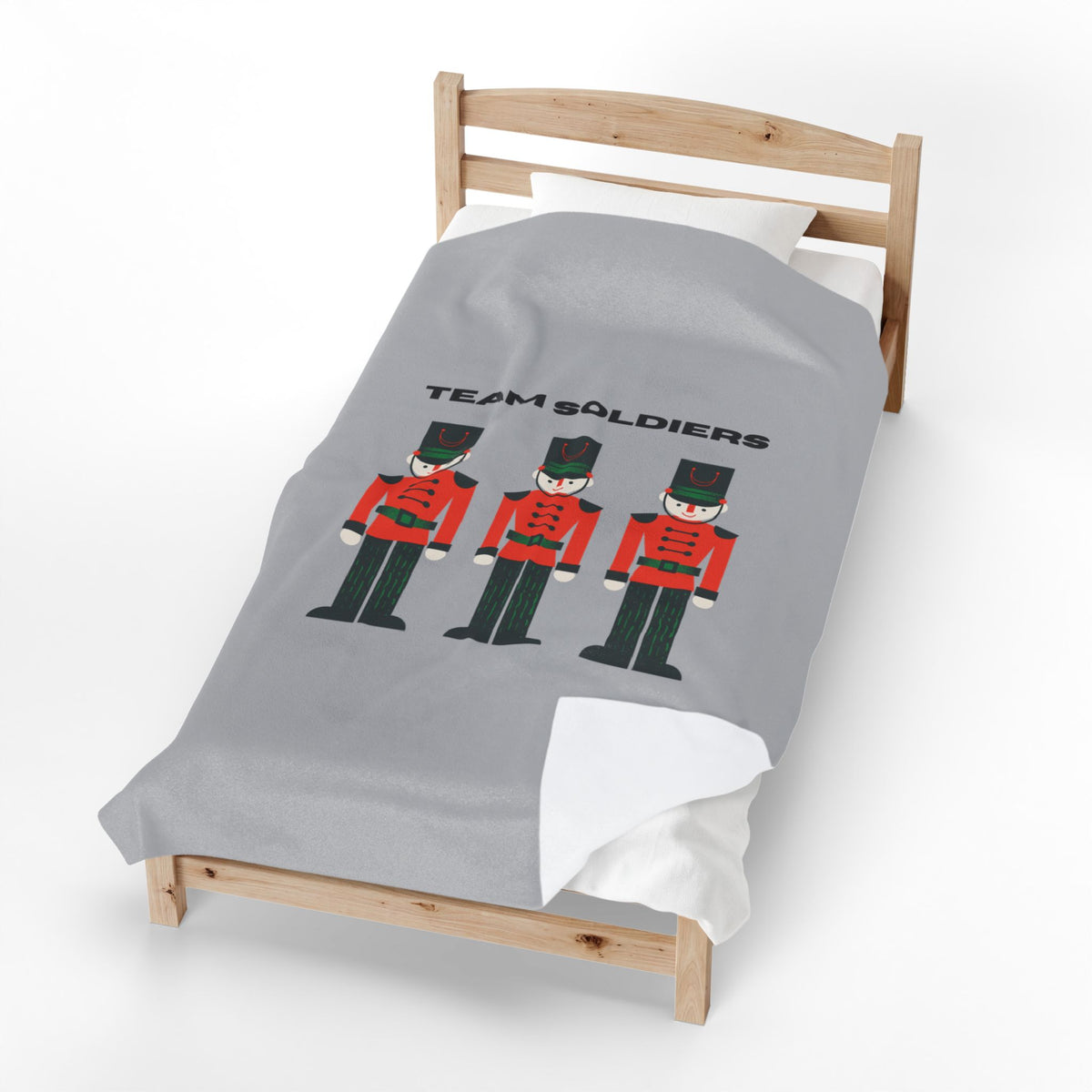 A light gray bedding set on a wooden toddler bed, featuring a graphic print of three toy soldier nutcrackers and the text 'TEAM SOLDIERS'.