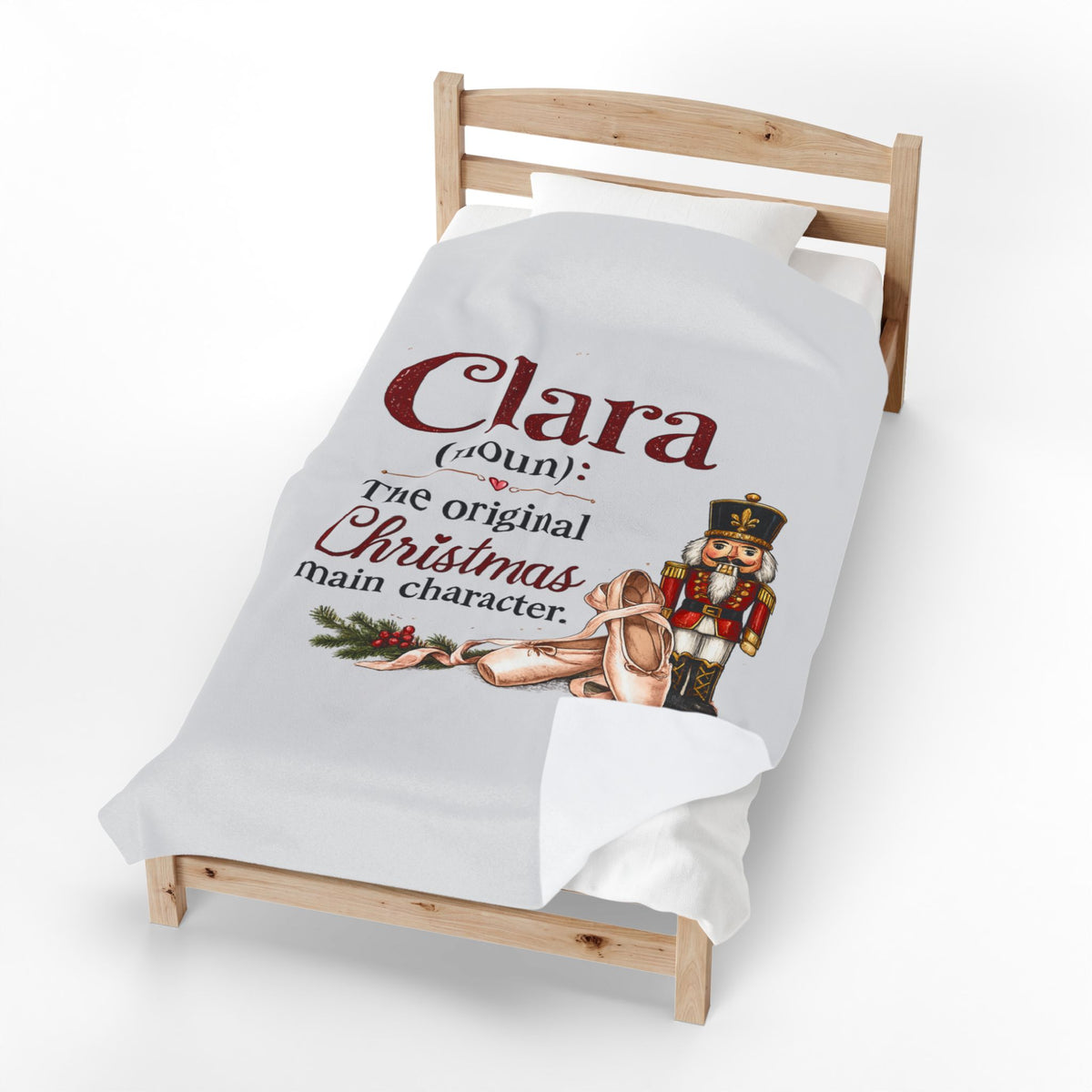 A white personalized Christmas blanket draped over a wooden toddler bed, featuring a printed design with the text 'Clara (Aunt) The original Christmas main character' alongside an illustration of a nutcracker and a ballerina.