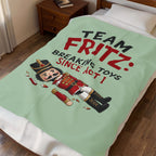 A mint green blanket with a cartoon nutcracker and the text 'TEAM FRITZ: BREAKING TOYS SINCE ACT I' spread across a bed in a bedroom setting.