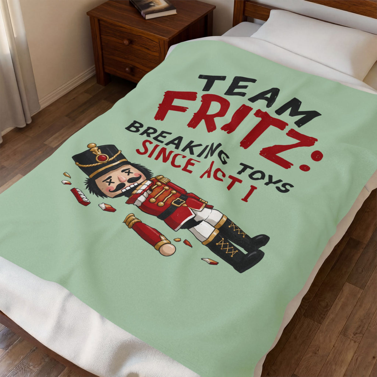 A mint green blanket with a cartoon nutcracker and the text 'TEAM FRITZ: BREAKING TOYS SINCE ACT I' spread across a bed in a bedroom setting.
