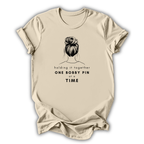 Beige t-shirt with a graphic of a bun and text 'holding it together ONE BOBBY PIN at a TIME' on a white background