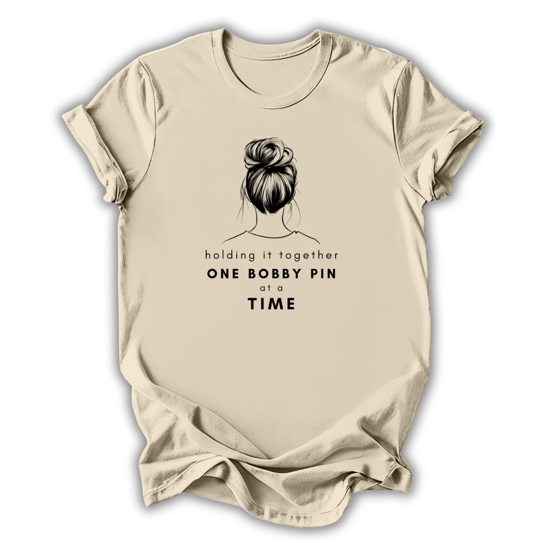 Beige t-shirt with a graphic of a bun and text 'holding it together ONE BOBBY PIN at a TIME' on a white background