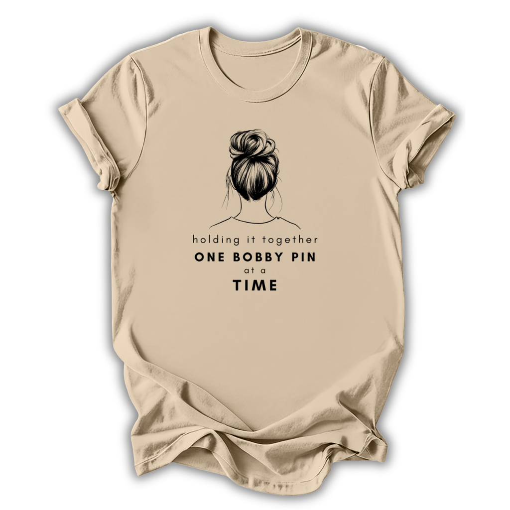 Beige t-shirt with graphic of a bun and text 'holding it together ONE BOBBY PIN AT A TIME' on a white background