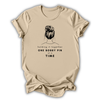 Beige t-shirt with graphic of a bun and text 'holding it together ONE BOBBY PIN AT A TIME' on a white background