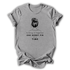 Gray t-shirt with graphic of a bun and text 'holding it together ONE BOBBY PIN AT A TIME' on a white background