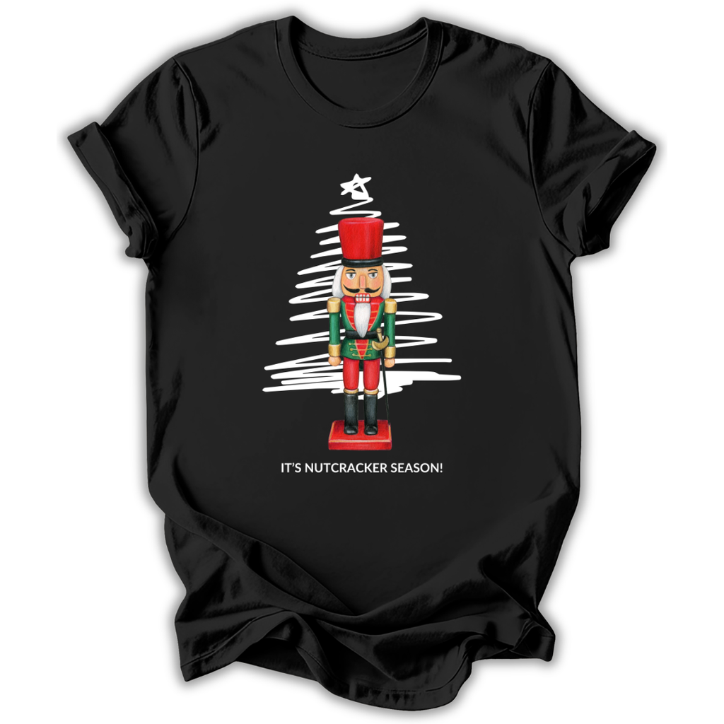 A black short-sleeve t-shirt with a graphic of a nutcracker standing in front of a white scribbled Christmas tree, with the text 'IT'S NUTCRACKER SEASON!' printed below.