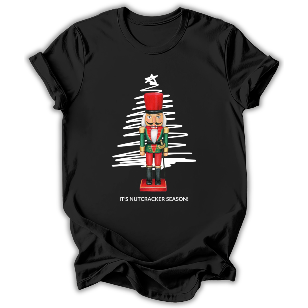 A black short-sleeve t-shirt with a graphic of a nutcracker standing in front of a white scribbled Christmas tree, with the text 'IT'S NUTCRACKER SEASON!' printed below.