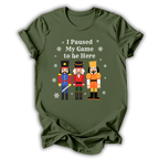 An olive green short-sleeve t-shirt with a graphic print of three nutcracker figures and the text 'I Paused My Game to Be Here' in white, displayed on a clean white background.
