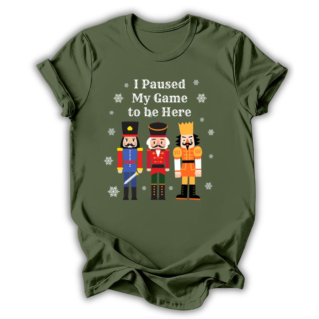 An olive green short-sleeve t-shirt with a graphic print of three nutcracker figures and the text 'I Paused My Game to Be Here' in white, displayed on a clean white background.