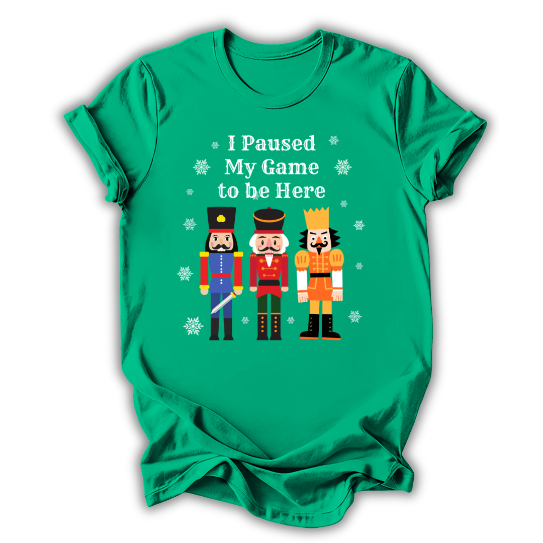 A green short-sleeve t-shirt with a graphic print of three nutcracker figures and the text 'I Paused My Game to be Here' in white, displayed on a clean white background.