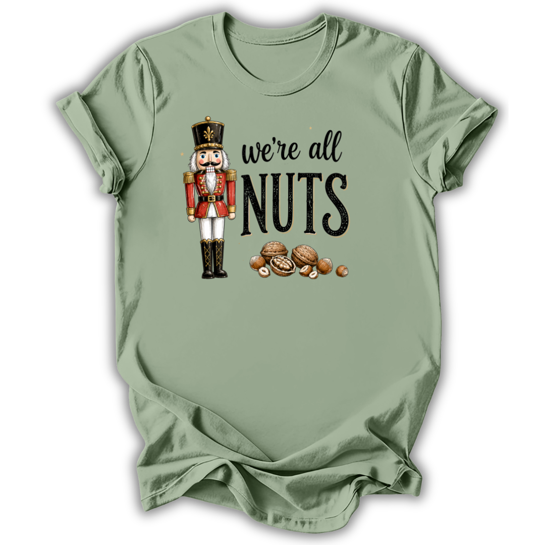 A sage green short-sleeve t-shirt with a graphic print of a nutcracker, the text 'we're all NUTS' in black, and several illustrated walnuts, displayed on a clean white background.