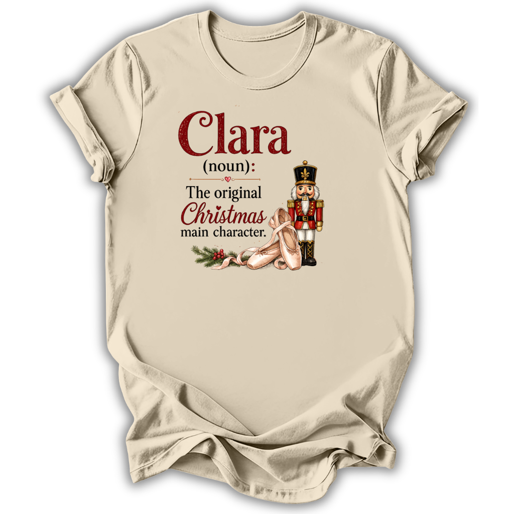 A beige short-sleeve t-shirt with a holiday-themed graphic print. The print features the text 'Clara (noun): The original Christmas main character.