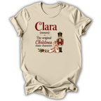 A beige short-sleeve t-shirt with a holiday-themed graphic print. The print features the text 'Clara (noun): The original Christmas main character.