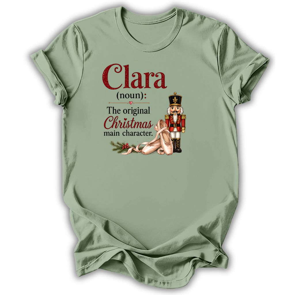 A sage green short-sleeve t-shirt with a humorous Christmas definition graphic printed on the front. The graphic reads 'Clara (noun): The original Christmas main character.