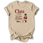 A beige short-sleeve t-shirt with a holiday-themed graphic print. The print features the text 'Clara (noun): The original Christmas main character.