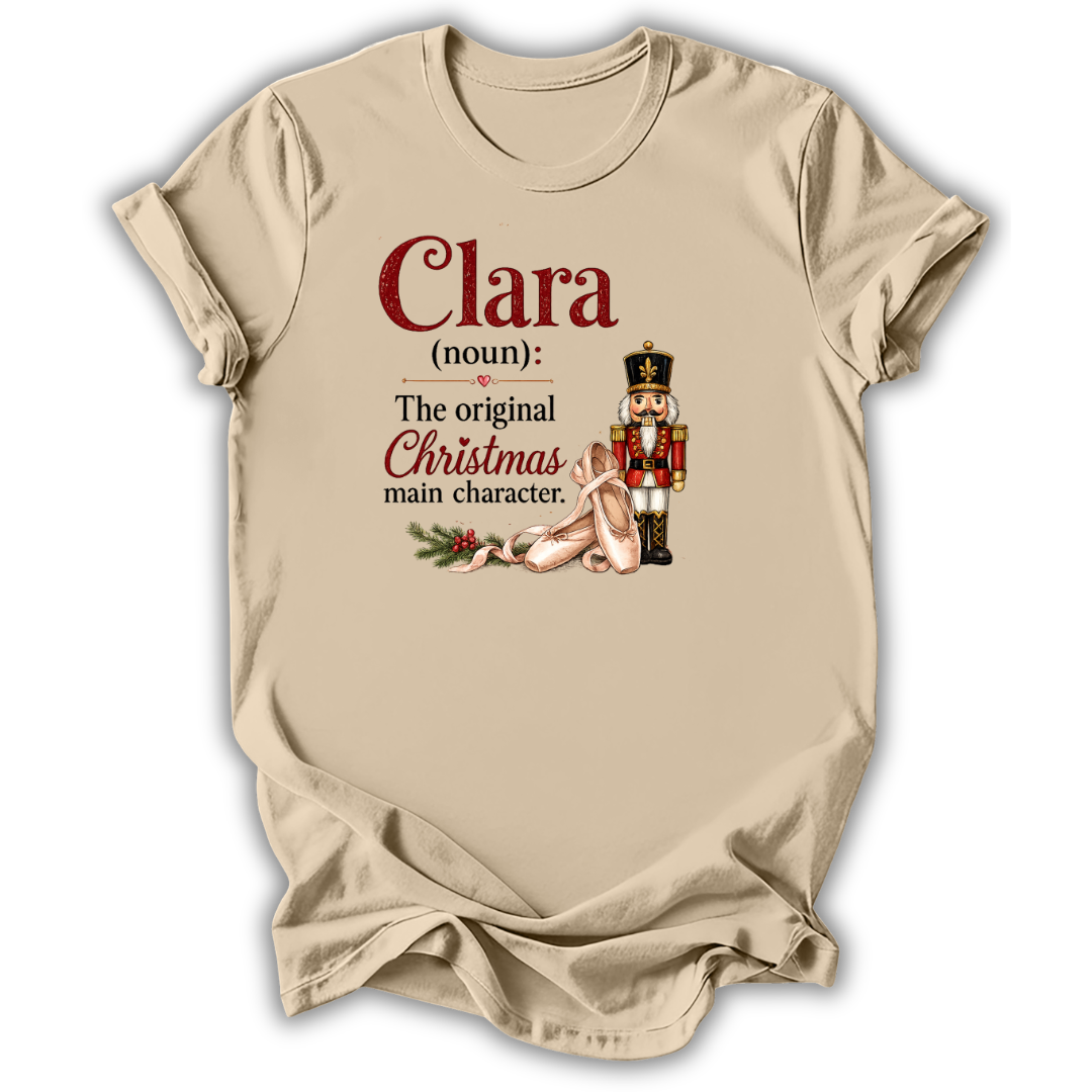 A beige short-sleeve t-shirt with a holiday-themed graphic print. The print features the text 'Clara (noun): The original Christmas main character.