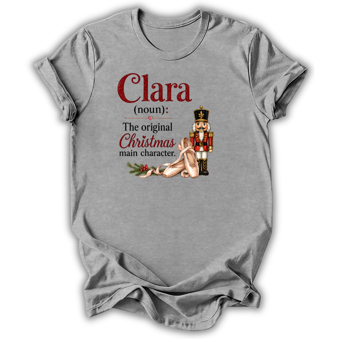 A heather gray short-sleeve t-shirt with a graphic print on the front. The print features the text 'Clara (noun): The original Christmas main character.