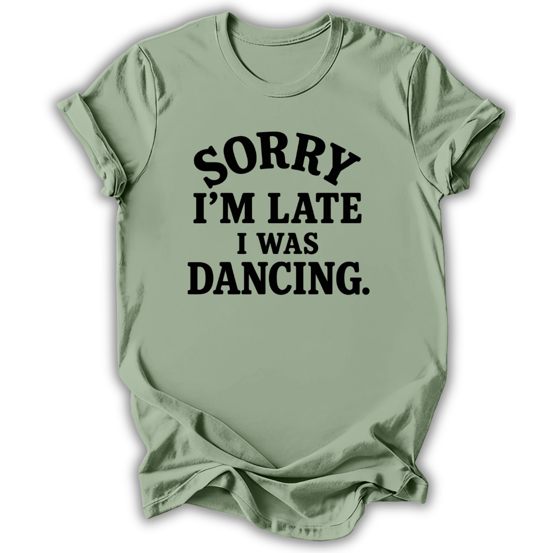 A sage green short-sleeve t-shirt with black text reading 'SORRY I'M LATE I WAS DANCING.' printed on the front, displayed against a white background.