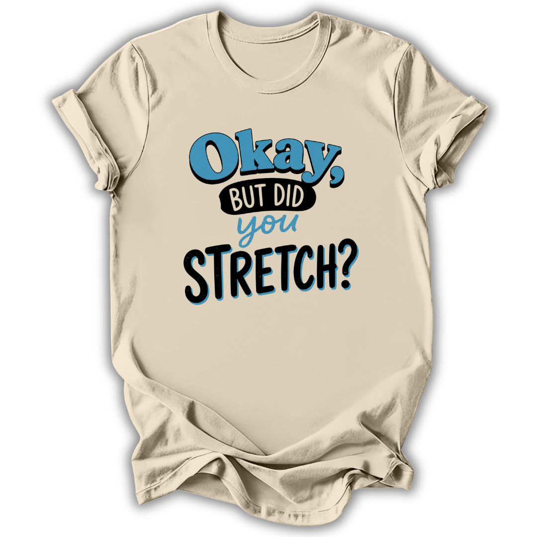 A beige short-sleeve t-shirt with a graphic print that reads 'Okay, BUT DID you STRETCH?' in blue and black lettering, displayed against a white background with a soft drop shadow.