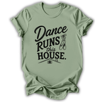 A sage green short-sleeve t-shirt with a black graphic print that reads 'Dance Runs This House.' accompanied by a ballet dancer ribbon bow and a small house illustration, displayed on a clean white background.
