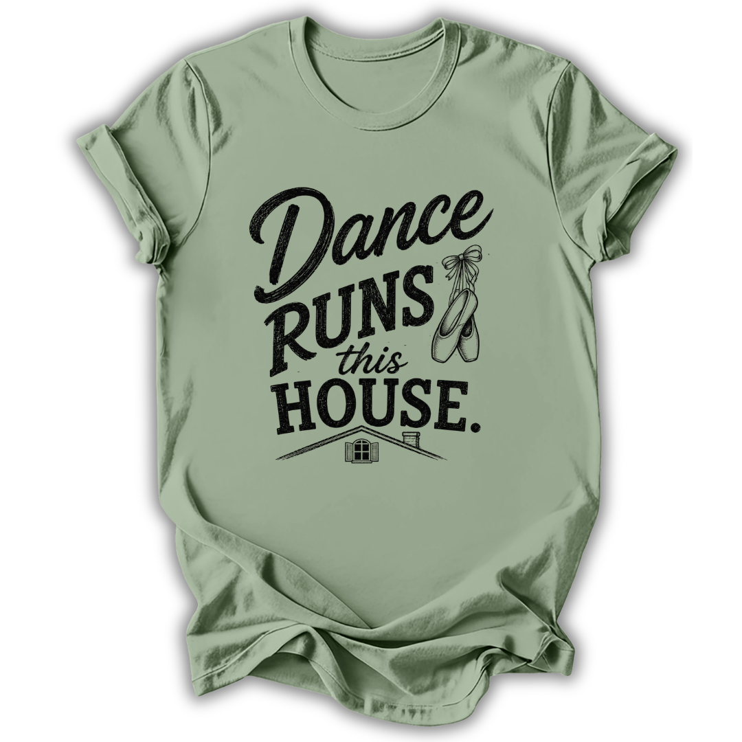 A sage green short-sleeve t-shirt with a black graphic print that reads 'Dance Runs This House.' accompanied by a ballet dancer ribbon bow and a small house illustration, displayed on a clean white background.