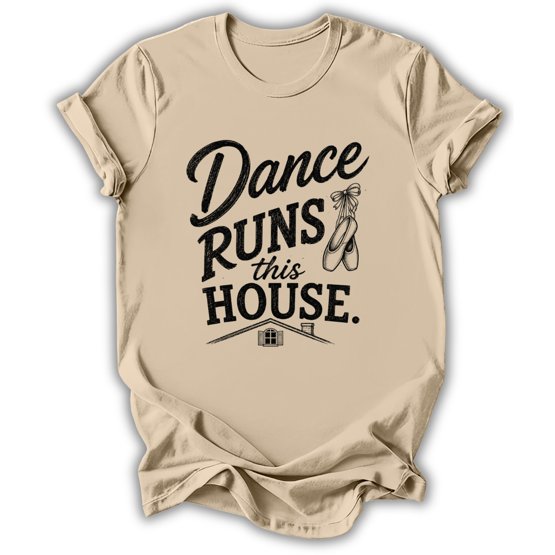 A beige short-sleeve t-shirt with a black graphic print that reads 'Dance Runs This House.' accompanied by ballet shoes and a small house illustration, displayed on a clean white background.