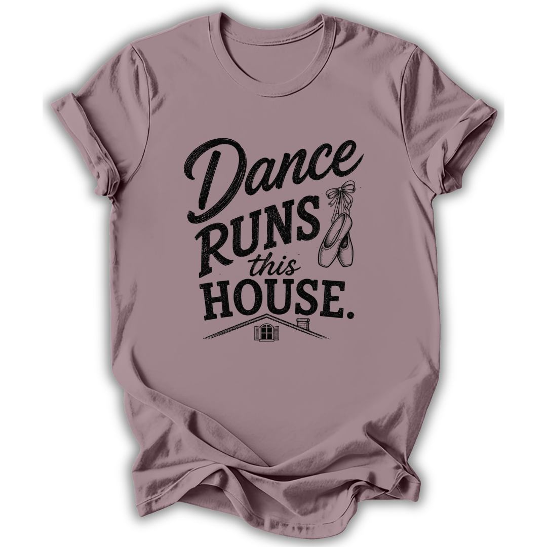 A dusty mauve short-sleeve t-shirt with a black graphic print that reads 'Dance Runs This House.' accompanied by a ballet dancer line drawing and a small house illustration, displayed on a clean white background.