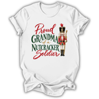 A white short-sleeve crewneck t-shirt with a graphic print that reads 'Proud GRANDMA of a NUTCRACKER Soldier' in red and green lettering, accompanied by an illustration of a nutcracker soldier.
