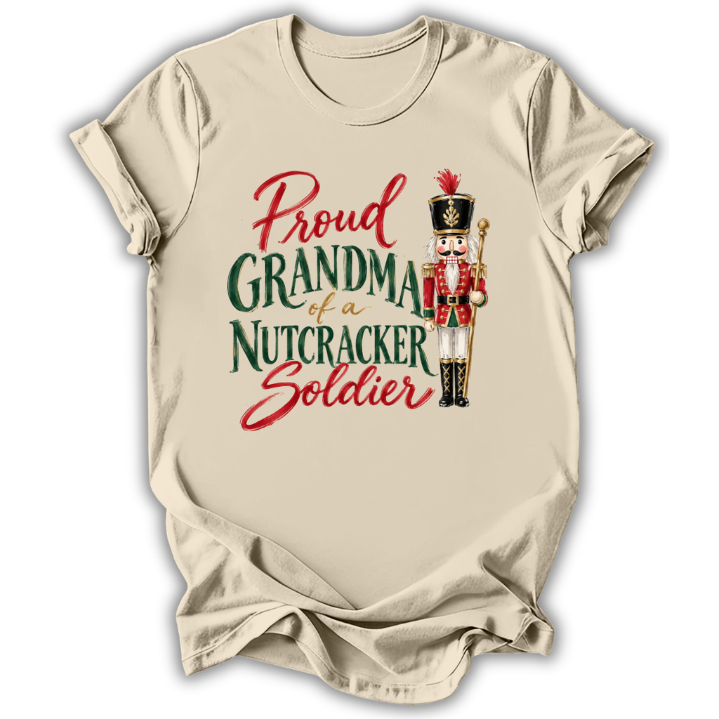 A beige short-sleeve t-shirt with a graphic print that reads 'Proud GRANDMA of a NUTCRACKER Soldier' in red and green lettering, accompanied by an illustration of a nutcracker soldier.