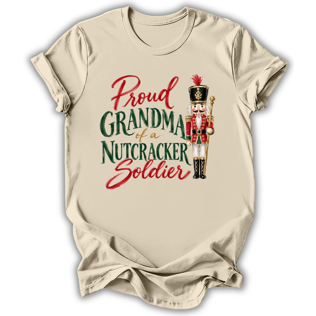 A beige short-sleeve t-shirt with a graphic print that reads 'Proud GRANDMA of a NUTCRACKER Soldier' in red and green lettering, accompanied by an illustration of a nutcracker soldier.