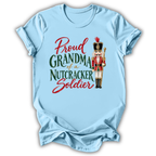 A light blue short-sleeve t-shirt with a graphic print that reads 'Proud GRANDMA of a NUTCRACKER Soldier' in red and green lettering, accompanied by an illustration of a nutcracker soldier, shown on a plain white background.