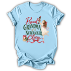 A light blue short-sleeve t-shirt with a vintage-style graphic on the front. The graphic features the text 'Proud GRANDMA of a Nutcracker Clara' in red and green lettering, accompanied by an illustration of a young girl in a white dress holding a nutcracker and some wrapped gifts.