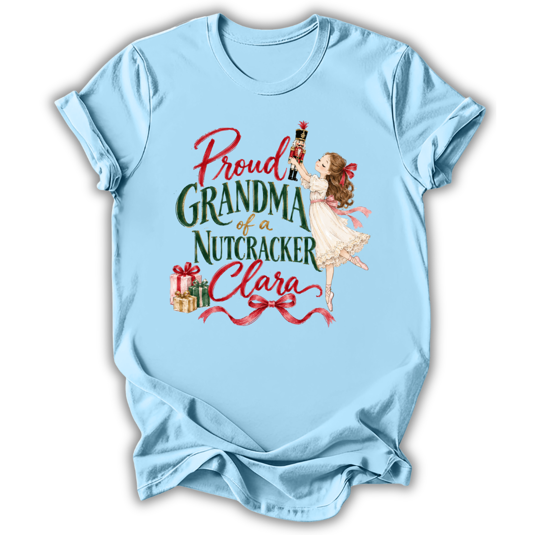 A light blue short-sleeve t-shirt with a vintage-style graphic on the front. The graphic features the text 'Proud GRANDMA of a Nutcracker Clara' in red and green lettering, accompanied by an illustration of a young girl in a white dress holding a nutcracker and some wrapped gifts.