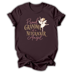 A dark plum-colored short-sleeve t-shirt with a graphic print on the front. The graphic features the text 'Proud GRANDMA of a NUTCRACKER Angel' in a mix of dusty pink and metallic gold fonts, accompanied by an illustration of a nutcracker ballerina in a white tutu.