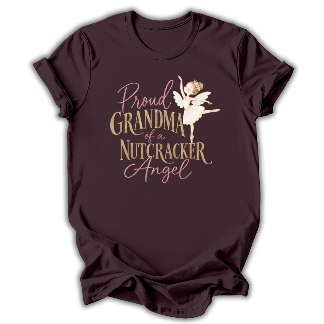 A dark plum-colored short-sleeve t-shirt with a graphic print on the front. The graphic features the text 'Proud GRANDMA of a NUTCRACKER Angel' in a mix of dusty pink and metallic gold fonts, accompanied by an illustration of a nutcracker ballerina in a white tutu.