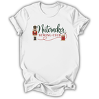 A white short-sleeve crewneck t-shirt with a 'Nutcracker Sewing Club' graphic on the chest, featuring a nutcracker soldier, green script text, and red embroidery details, shown on a white background.