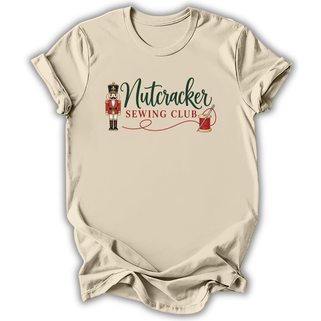 A beige short-sleeve crewneck t-shirt with a graphic on the chest that reads 'Nutcracker Sewing Club' in green and red script, accompanied by a nutcracker figure and a red spool of thread, shown on a white background.
