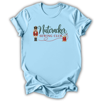 A light blue short-sleeve crewneck t-shirt with a graphic on the chest that reads 'Nutcracker Sewing Club' in dark green and red lettering, accompanied by an illustration of a nutcracker and a spool of thread.
