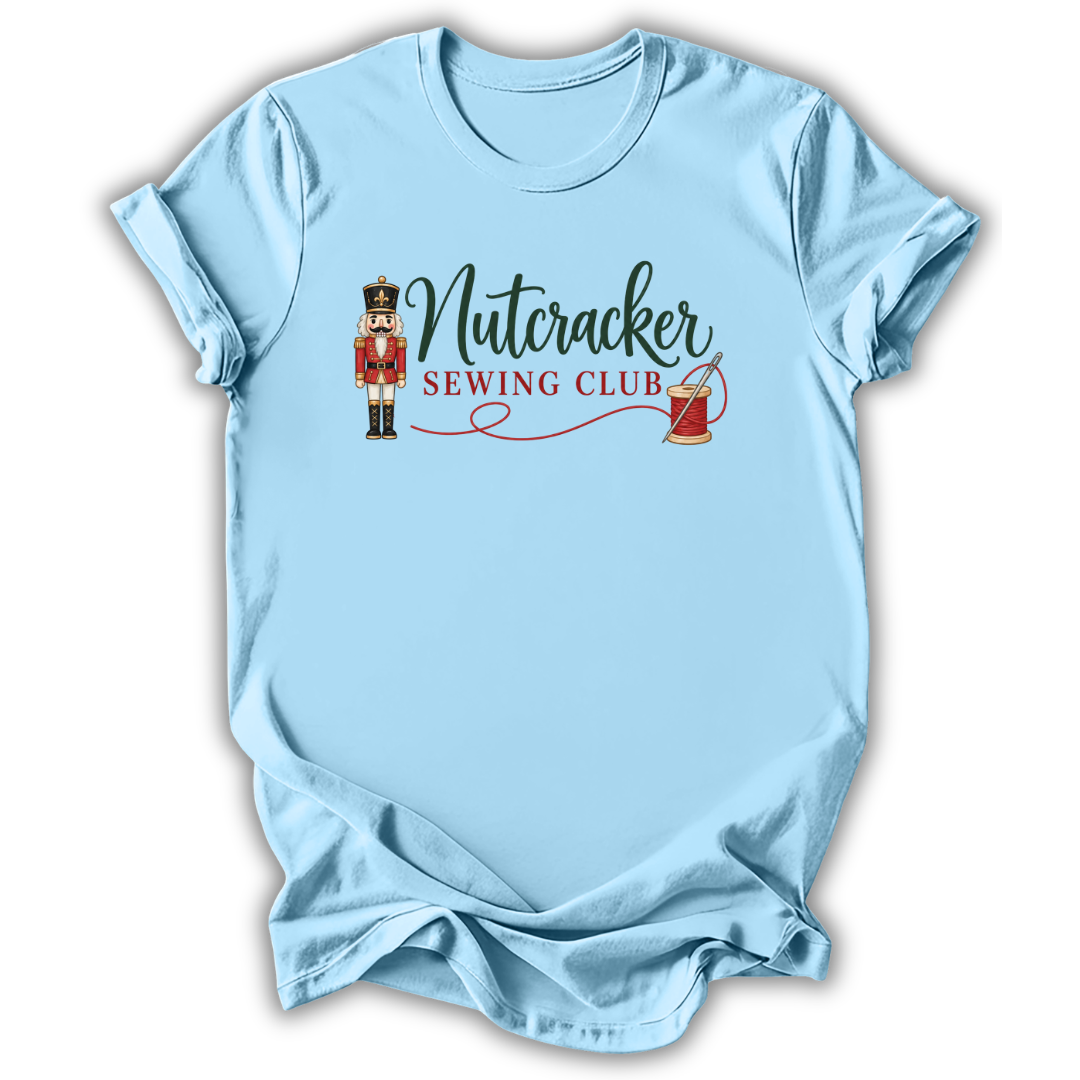 A light blue short-sleeve crewneck t-shirt with a graphic on the chest that reads 'Nutcracker Sewing Club' in dark green and red lettering, accompanied by an illustration of a nutcracker and a spool of thread.