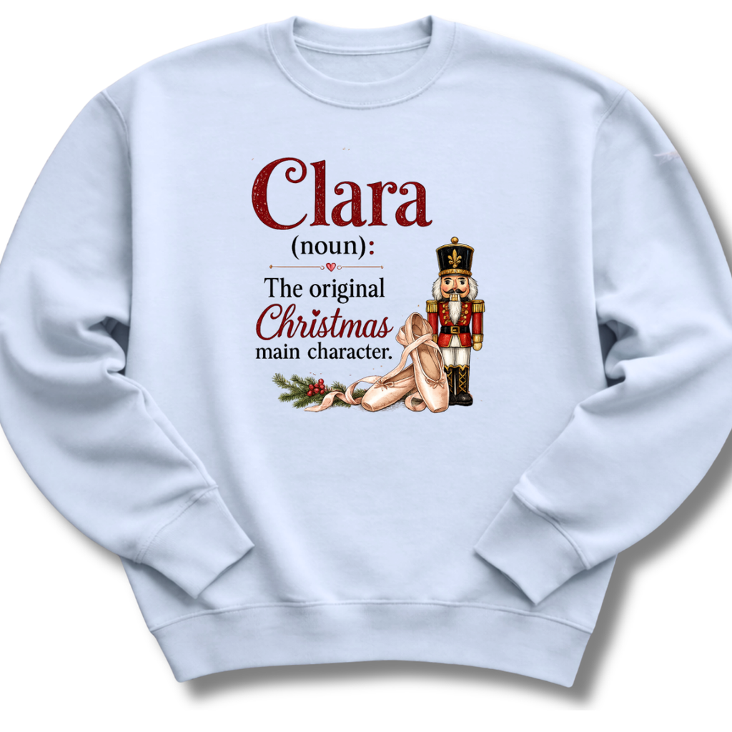 A light blue crewneck sweatshirt featuring a custom Christmas-themed graphic print on the front. The graphic defines the name 'Clara' as 'The original Christmas main character' with an illustration of a sleeping nutcracker and a sack of coins.