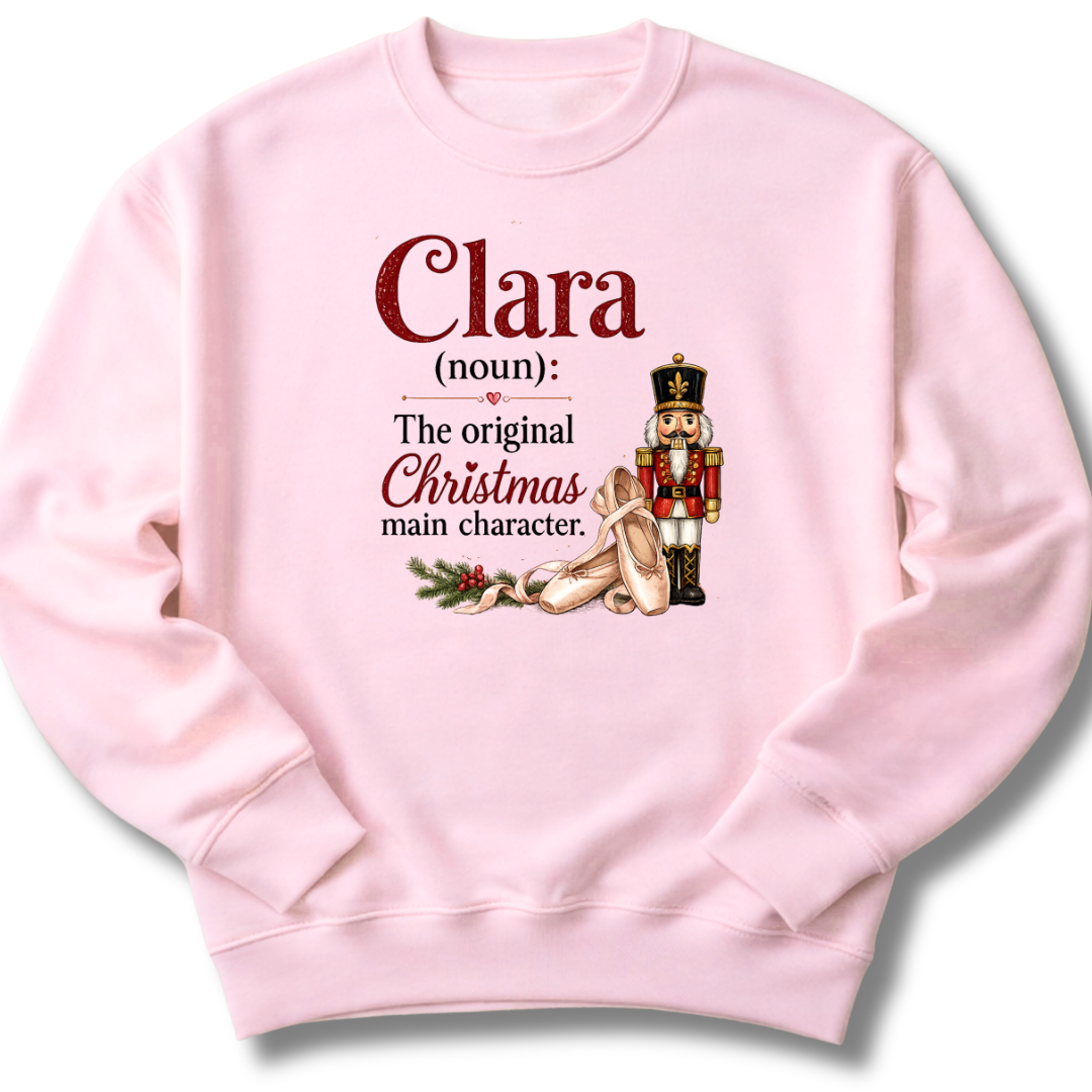 A light pink crewneck sweatshirt featuring a custom holiday graphic with the text 'Clara (noun): The original Christmas main character.' and an illustration of a nutcracker and ballet shoes.